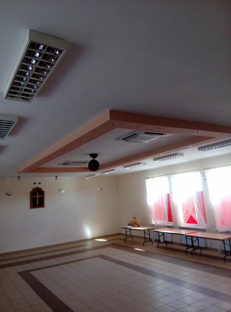 Wnętrze jasnego pomieszczenia z kasetonowymi klimatyzatorami w podwieszanym suficie z dekoracyjną ramą, widoczne także lampy rastrowe i wiszący wentylator sufitowy.