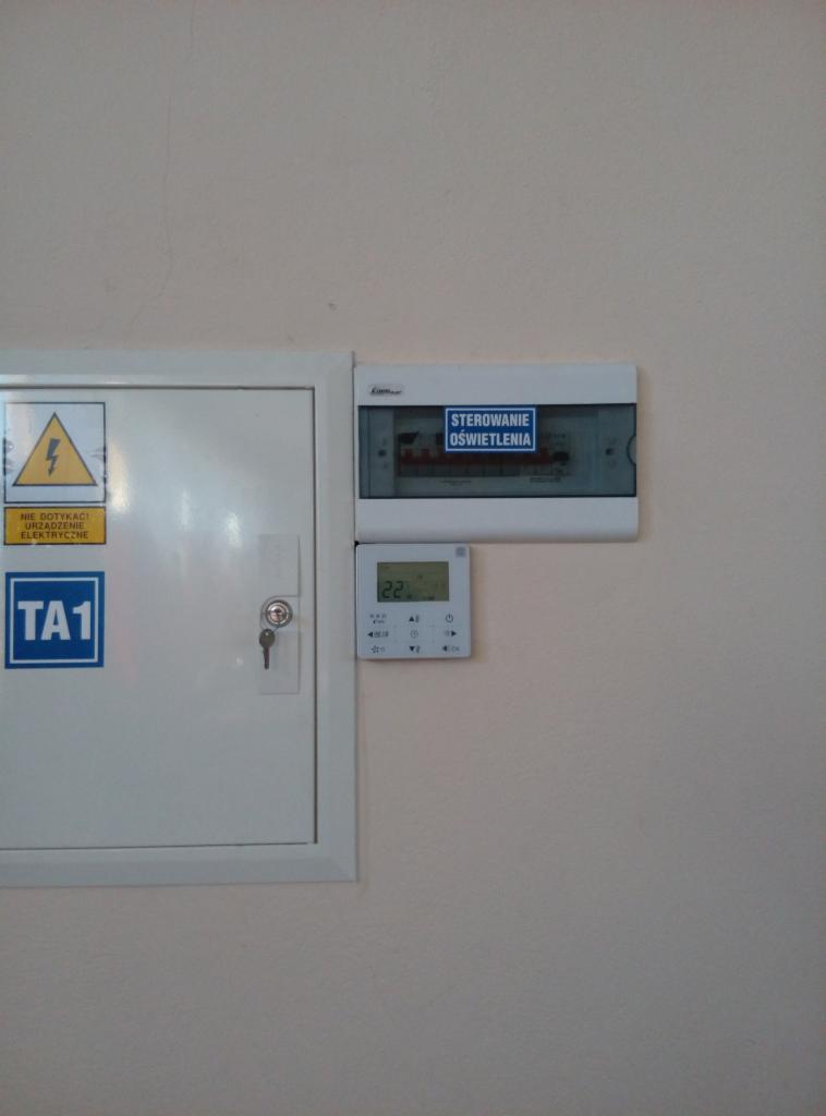Panel sterowania oświetleniem i termostat na ścianie, obok skrzynki elektrycznej z ostrzeżeniem o napięciu.