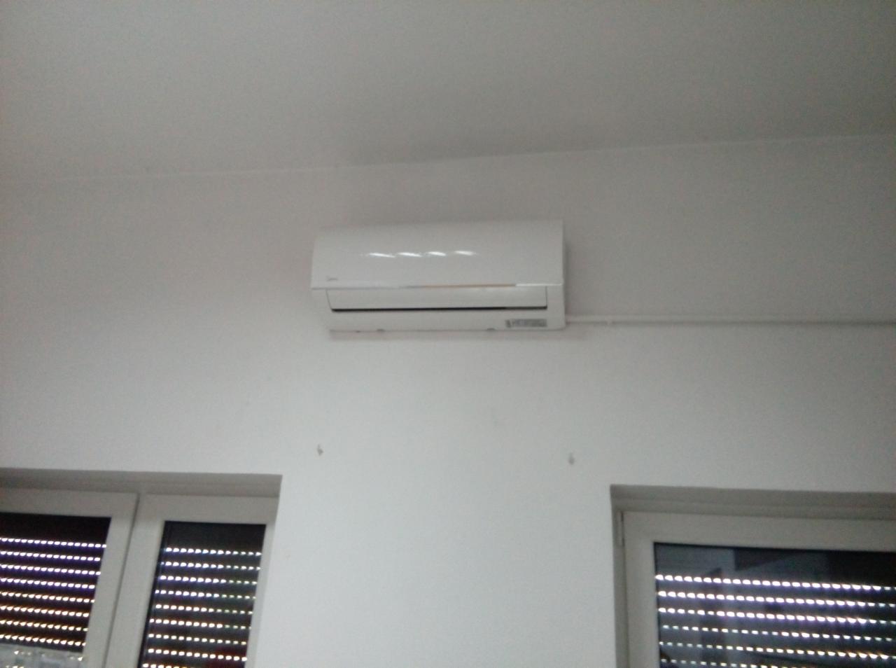 Biały klimatyzator ścienny Midea zamontowany w biurze nad oknami z żaluzjami, widoczne rurki instalacyjne.