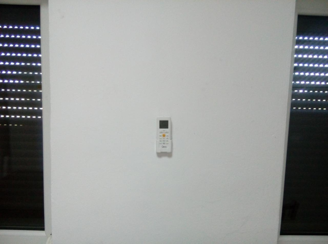 Biała ściana z zamontowanym pilotem do klimatyzacji marki Midea, widoczne rolety okienne.