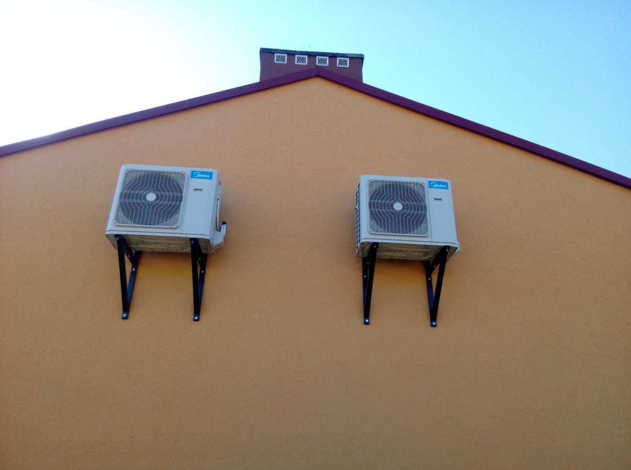Dwa identyczne klimatyzatory Midea zamontowane na czarnych wspornikach na pomarańczowej ścianie budynku z ciemnoczerwonym dachem i kominem na tle błękitnego nieba.