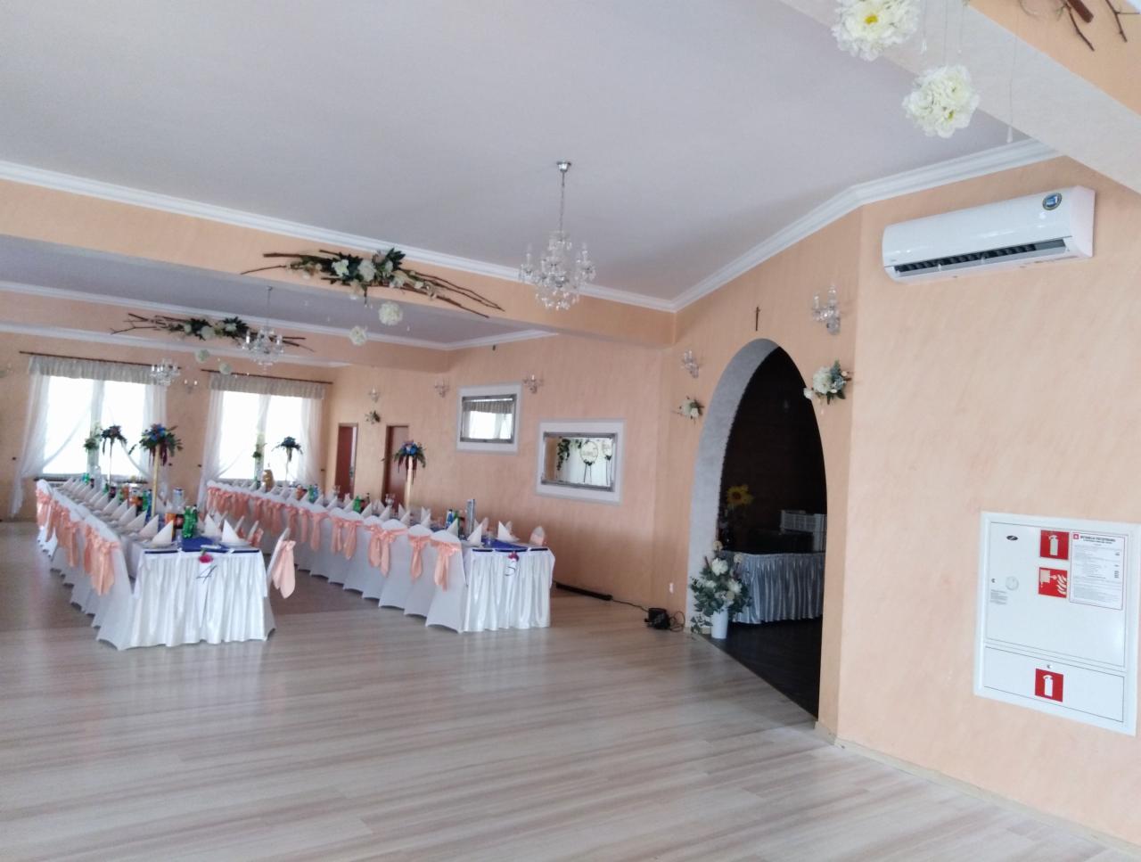 Sala bankietowa z długimi stołami przygotowanymi do uroczystości, klimatyzator ścienny w prawym górnym rogu, dekoracje kwiatowe i żyrandole