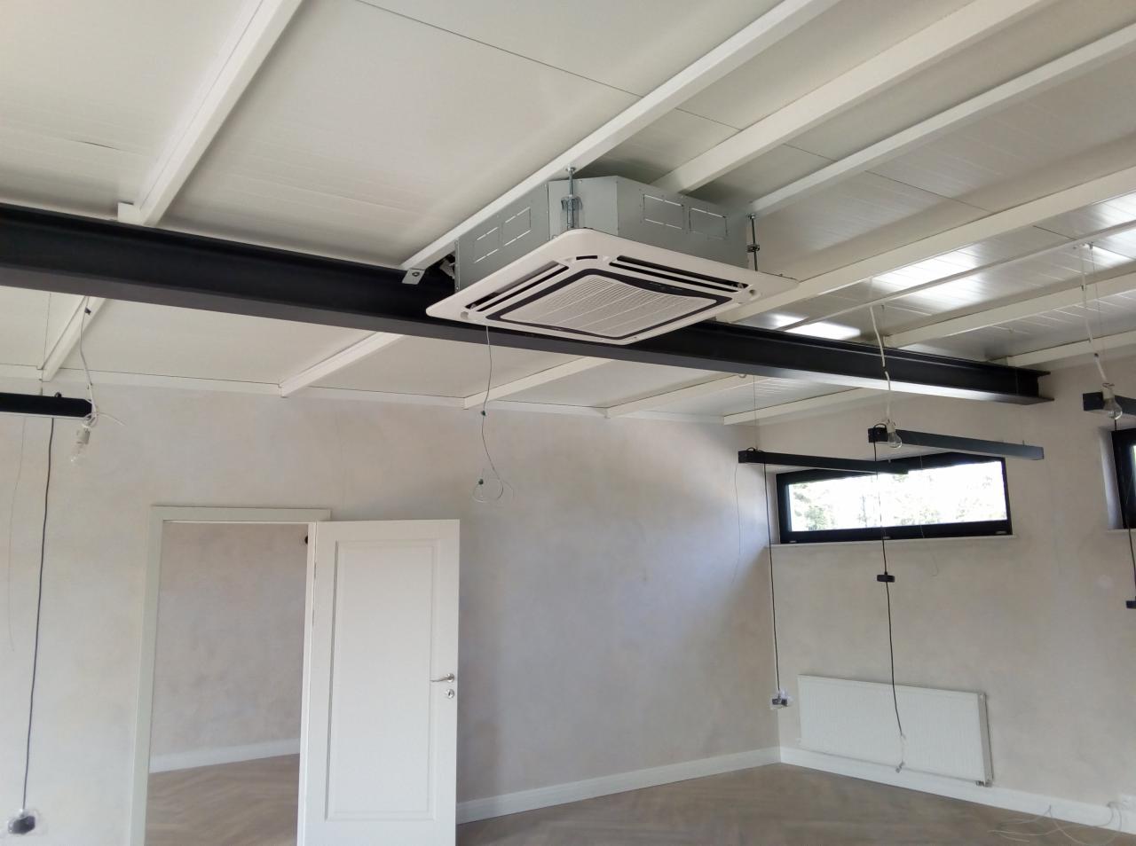 Kasetonowa jednostka klimatyzacyjna zamontowana pod sufitem w pomieszczeniu biurowym z widocznymi elementami konstrukcyjnymi sufitu i oknami.