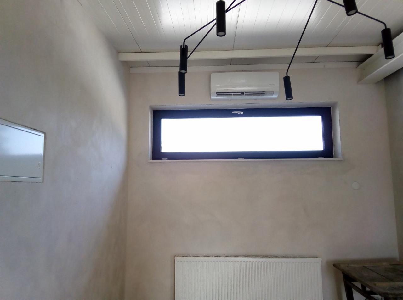 Biała jednostka klimatyzacyjna zamontowana nad oknem w pomieszczeniu z kremowymi ścianami, białym sufitem i nowoczesną czarną lampą sufitową.