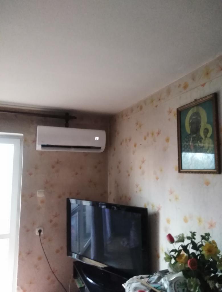 Biała jednostka klimatyzacyjna zamontowana na ścianie w pokoju z kwiecistą tapetą, widoczny również telewizor i obraz na ścianie.