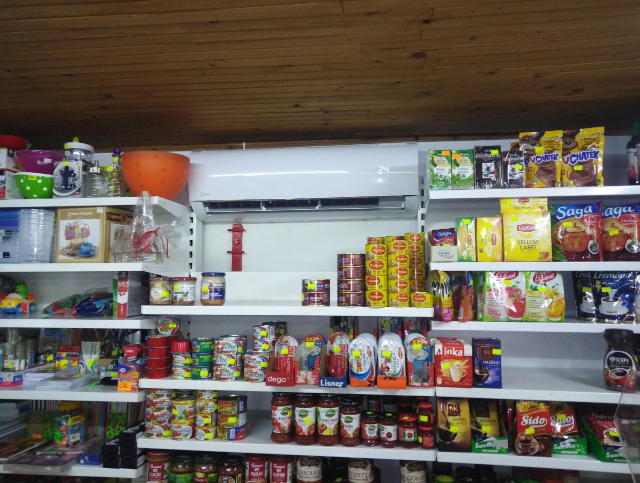 Biały klimatyzator ścienny zamontowany w sklepie spożywczym, widoczny nad półkami z różnorodnymi produktami spożywczymi.