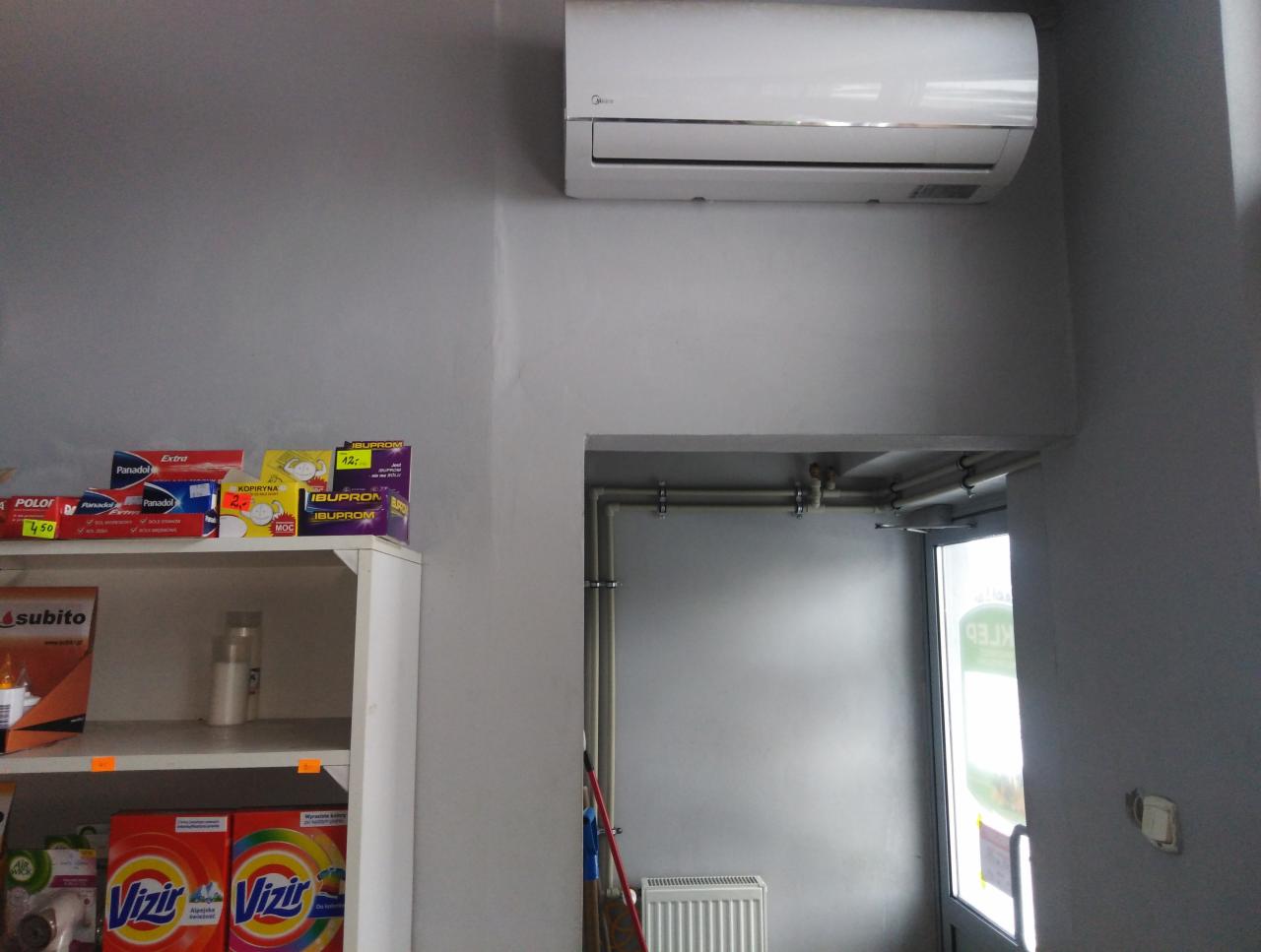 Biały klimatyzator Midea zamontowany nad wejściem do sklepu, widoczny regał z produktami spożywczymi i chemicznymi, szare ściany.