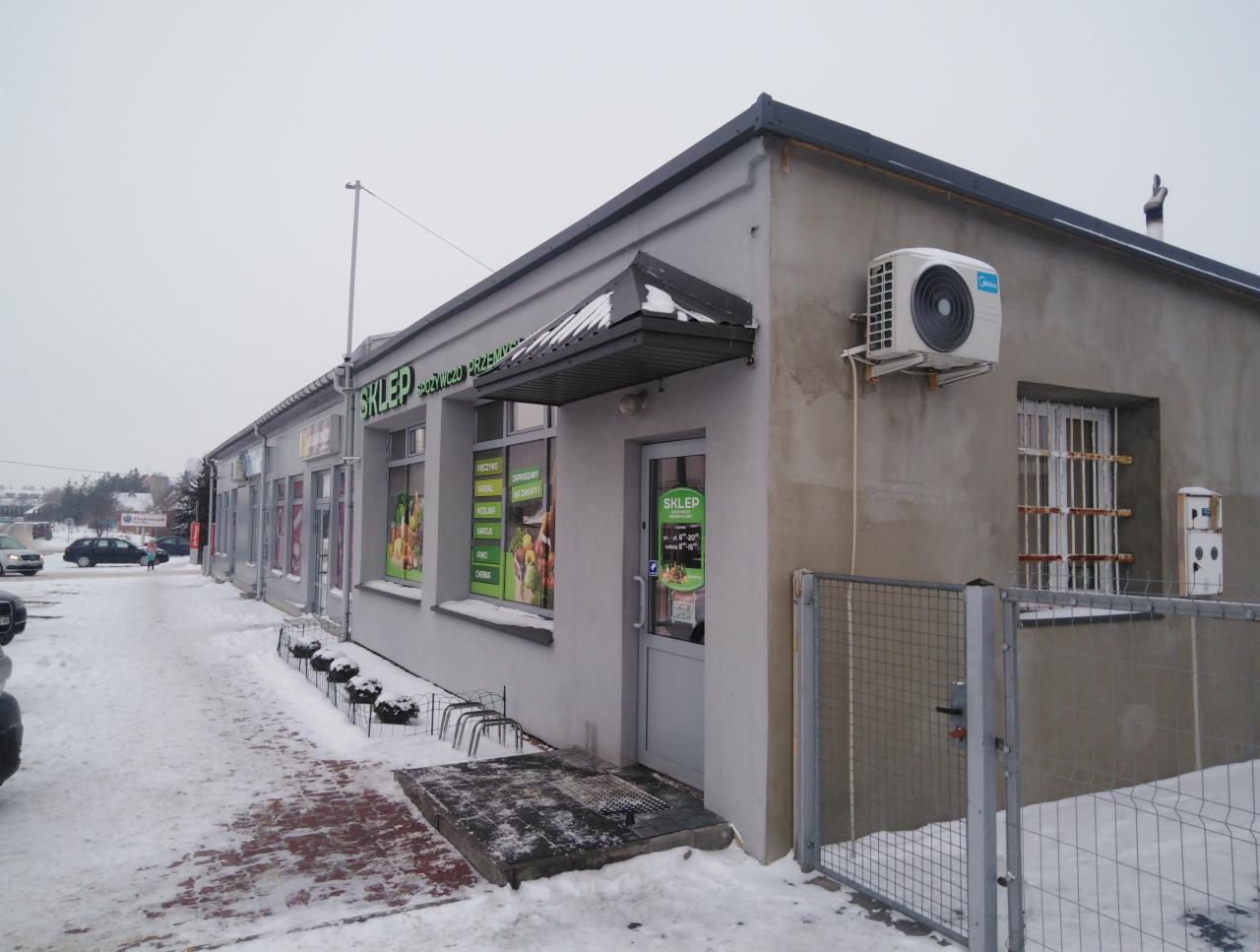 Zewnętrzna jednostka klimatyzacyjna Midea zamontowana na szarej ścianie sklepu spożywczo-przemysłowego w zimowy dzień.