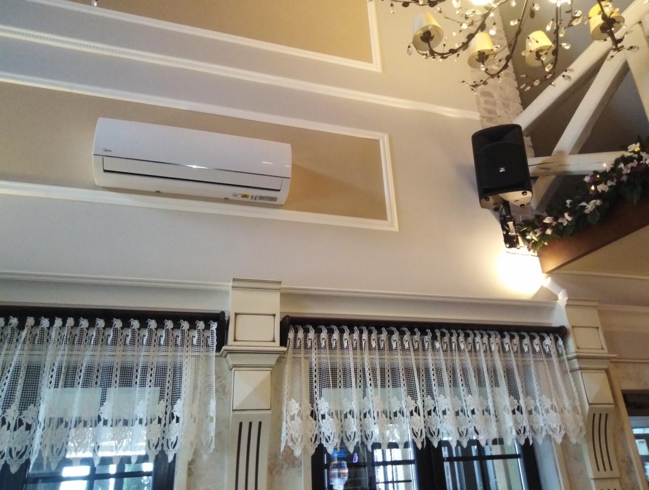 Biały klimatyzator Midea zamontowany pod sufitem w pomieszczeniu z dekoracyjnymi zasłonami i żyrandolem, widok z dołu.