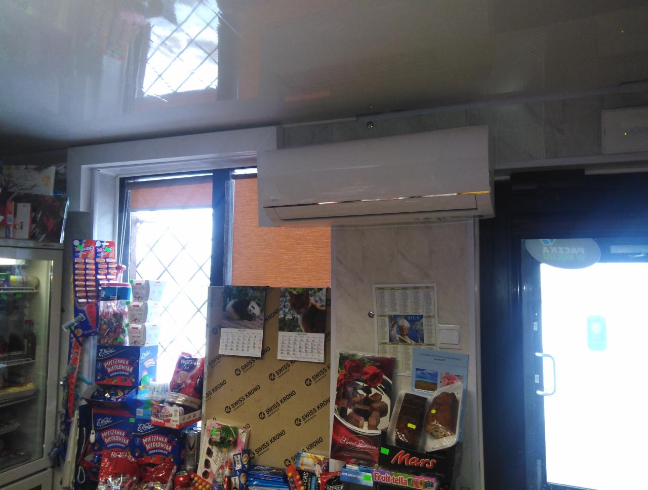 Zamontowany klimatyzator w sklepie spożywczym w Łosicach, widoczny nad regałami ze słodyczami i kalendarzami. Biała obudowa urządzenia kontrastuje z ciemnym tłem okna i drzwi.