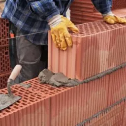 Murowanie ściany z czerwonych, porowatych cegieł, zaprawa murarska i kielnia w kadrze, widoczne fragmenty osoby w roboczych rękawicach.