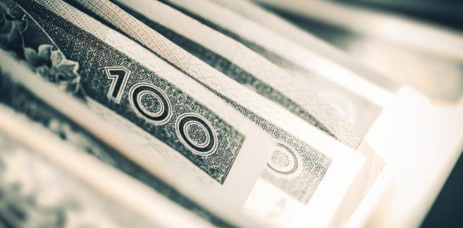 Stos banknotów 100 zł w odcieniach szarości, ułożonych warstwami z widocznymi fragmentami nominałów i zabezpieczeń.