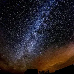 Nocne niebo usiane gwiazdami, z wyraźnie widoczną Drogą Mleczną, sfotografowane nad sylwetką domu i drzew.