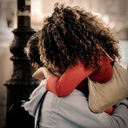 Dream come true s.r.o. - Para obejmuje się na tle miejskiego krajobrazu, ona w czerwonej bluzce i z torbą na ramieniu, on w jasnej bluzie z kapturem, widoczne kręcone włosy kobiety i fragment latarni