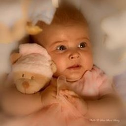 Dream come true s.r.o. - Urocze niemowlę w różowej sukience, leżące na plecach, przytulające pluszowego misia w pastelowych kolorach, z delikatnym rozmyciem tła i miękkim światłem.