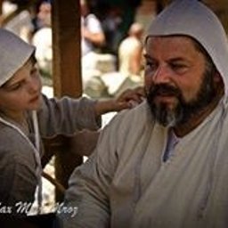 Dream come true s.r.o. - Chłopiec w szarym stroju i kapturze dotyka ramienia brodatego mężczyzny w jasnym stroju, obaj siedzą na ławce w plenerze podczas wydarzenia historycznego.