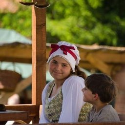 Dream come true s.r.o. - Dwoje dzieci w strojach historycznych siedzi przy drewnianym stole pod zadaszeniem, w tle zieleń i waga szalkowa.