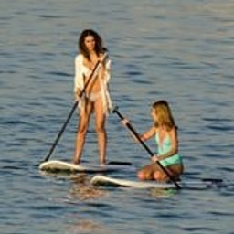 Dream come true s.r.o. - Dwie kobiety na deskach SUP na spokojnym morzu, jedna stoi w białej narzutce, druga klęczy w turkusowym stroju, obie trzymają wiosła.