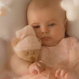 Dream come true s.r.o. - Niemowlę w różowej tiulowej sukience, tulące pluszowego misia w czapeczce, w łóżeczku z dekoracjami w pastelowych barwach.