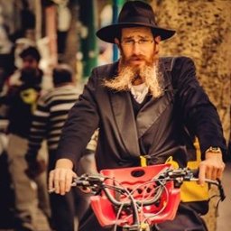 Dream come true s.r.o. - Portret brodatego mężczyzny w czarnym kapeluszu i płaszczu na rowerze z czerwonym koszykiem na tle ulicy.