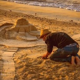 Dream come true s.r.o. - Mężczyzna w kapeluszu klęczy na plaży, tworząc skomplikowaną rzeźbę z piasku z motywem zamku i schodów, z falami morskimi w tle.