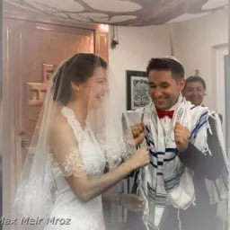 Uśmiechnięta para młoda podczas żydowskiego ślubu, wymiana obrączek pod baldachimem, mężczyzna w tałesie i czerwonej muszce.