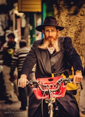 Portret brodatego mężczyzny w czarnym kapeluszu i płaszczu na rowerze z czerwonym koszykiem na tle ulicy.