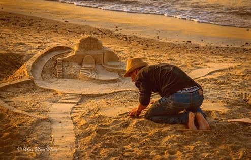 Mężczyzna w kapeluszu klęczy na plaży, tworząc skomplikowaną rzeźbę z piasku z motywem zamku i schodów, z falami morskimi w tle.