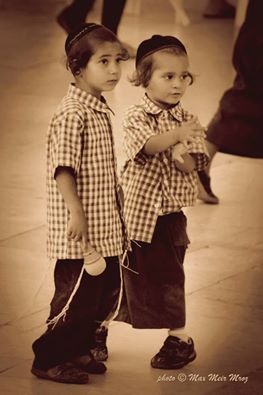 Dwaj mali chłopcy w jarmułkach i kraciastych koszulach, jeden trzyma zabawkę, patrzą w dal na sepiowym zdjęciu.