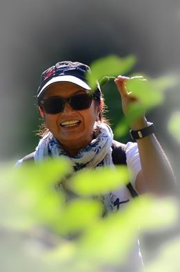 Uśmiechnięta kobieta w okularach przeciwsłonecznych i czapce z daszkiem trzyma liść na tle zieleni.