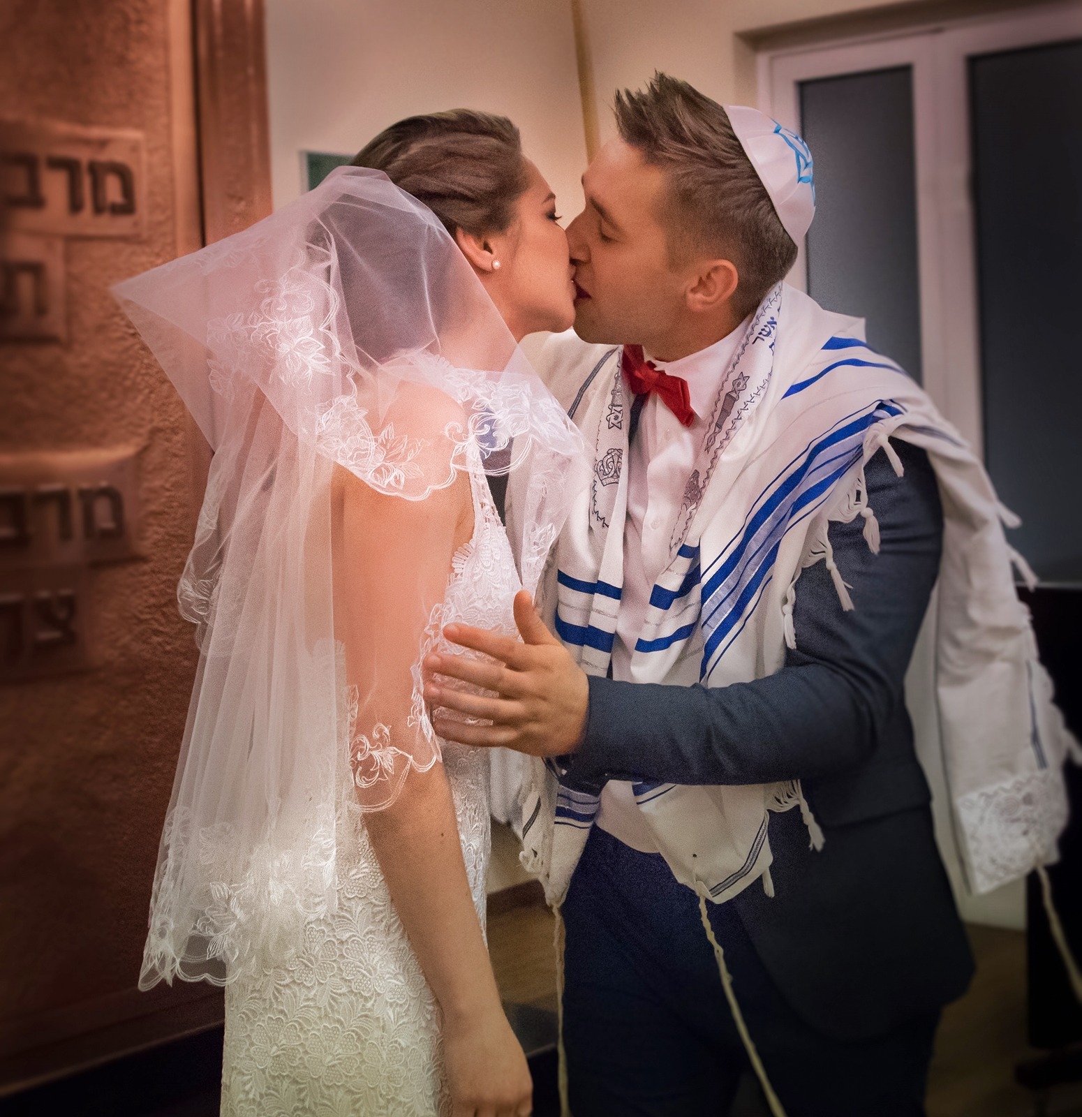 Pocałunek młodej pary pod ozdobnym baldachimem podczas ceremonii ślubnej w synagodze, mężczyzna w jarmułce i tałesie.