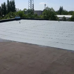 Płaski dach pokryty szarymi, prostokątnymi płytami bitumicznymi, widok z góry na tle zieleni drzew i budynków w oddali.