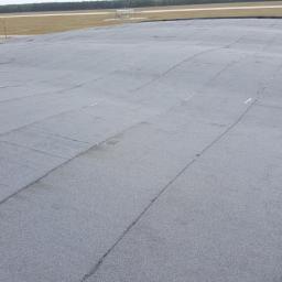 Usługi Dekarskie - Szary, płaski dach pokryty papą termozgrzewalną na obiekcie przemysłowym, widoczna linia horyzontu i fragment sylwetki pracownika w oddali.