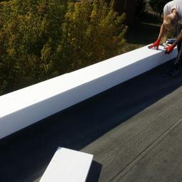 Usługi Dekarskie - Pracownik w czerwonych rękawicach montuje białe elementy izolacyjne na płaskim dachu, widok z góry, w tle drzewa.