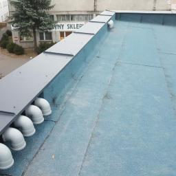 Usługi Dekarskie - Płaski dach pokryty papą w kolorze błękitnym, z szarymi obróbkami blacharskimi i systemem odprowadzania wody w postaci białych rur PCV, widok z góry na budynek z otoczeniem.