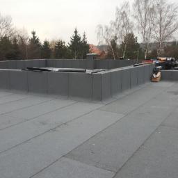 Usługi Dekarskie - Prace dekarskie na płaskim dachu pokrytym papą termozgrzewalną, dwóch dekarzy w trakcie łączenia papy za pomocą palnika, widok na drzewa w tle.