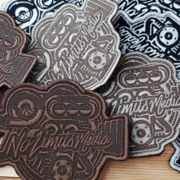 Kolekcja haftowanych naszywek z motywami motoryzacyjnymi i napisem 'No Limits Media' w kolorach brązowym, beżowym i czarno-białym, ułożonych na drewnianym tle.