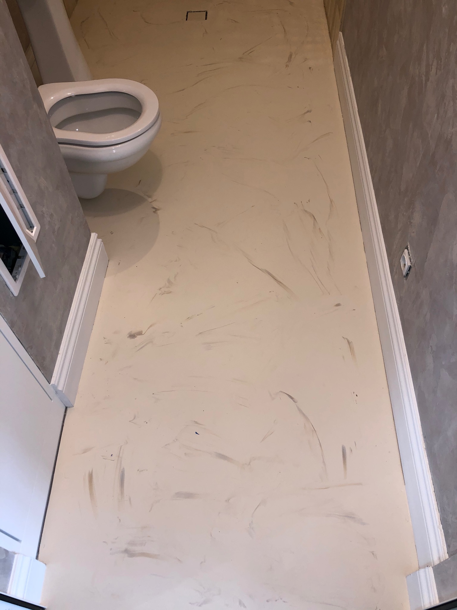 Beżowa posadzka żywiczna z szarymi smugami w małej łazience z toaletą, widoczny fragment ściany z listwą przypodłogową.
