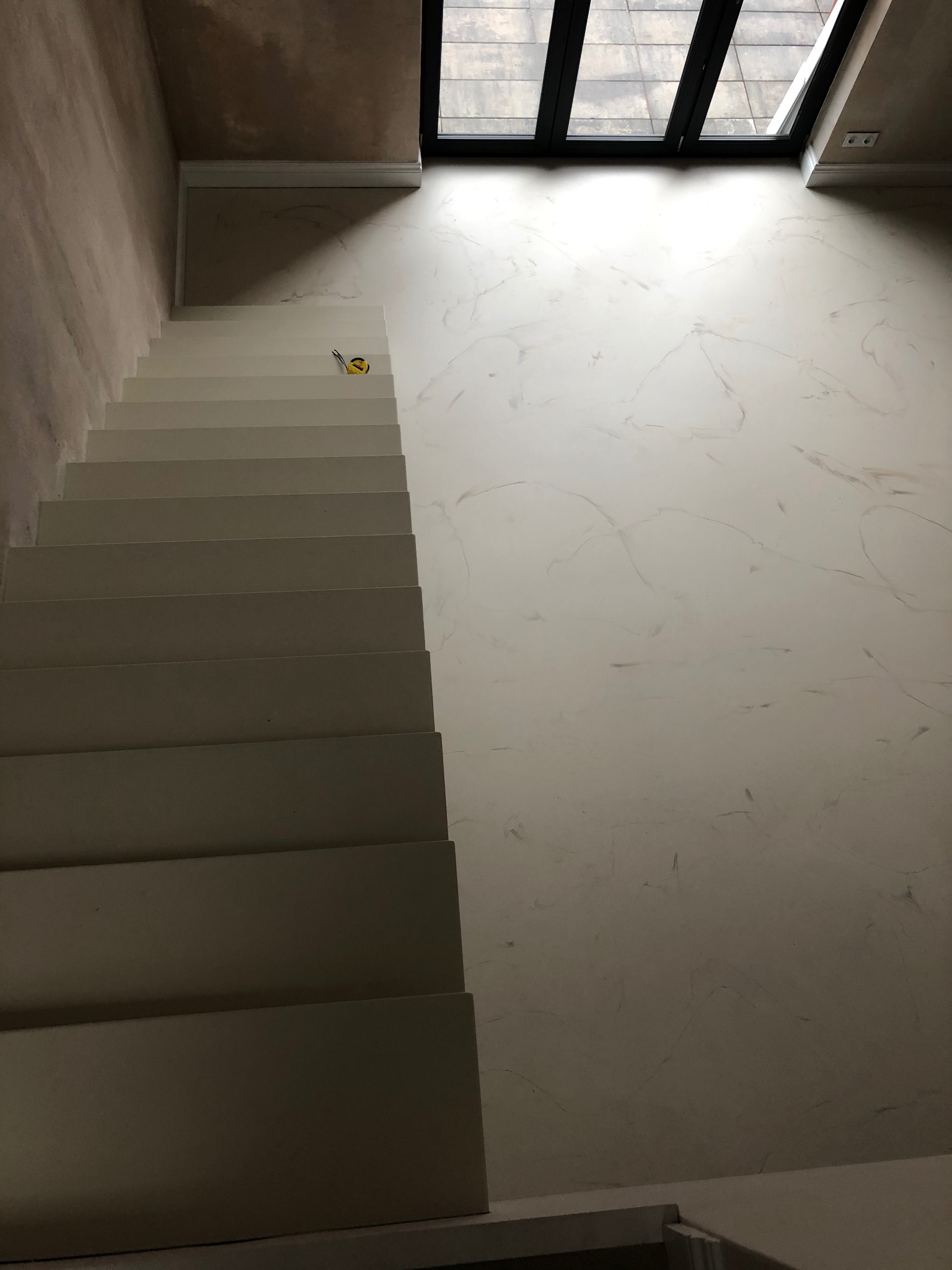 Widok z góry na białe schody z taśmą mierniczą leżącą na jednym ze stopni, ściana wykończona w stylu marmuru, szklany dach w tle.
