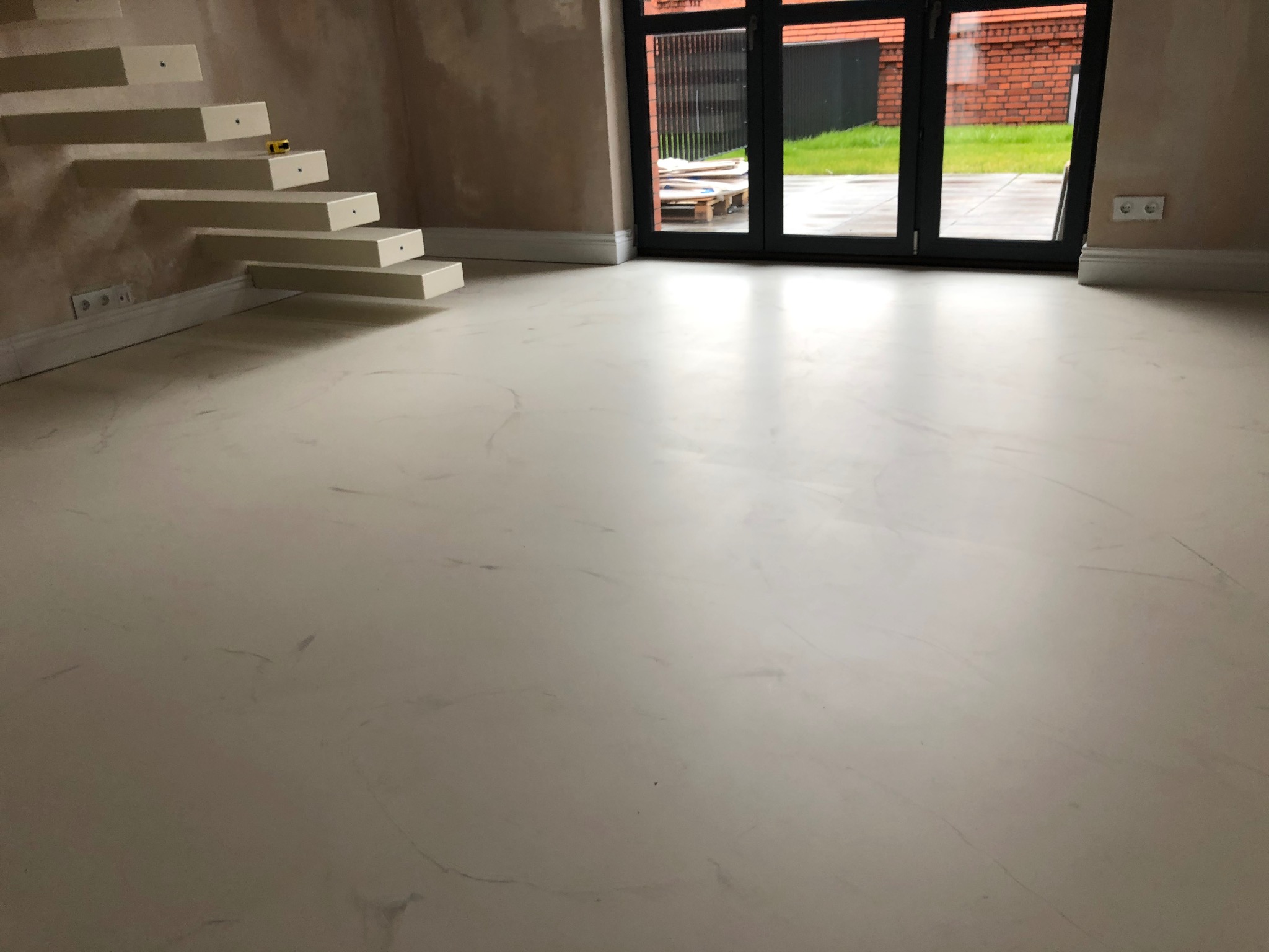 Jasnoszara podłoga z delikatnym wzorem marmuru, schody wiszące i duże okno balkonowe z widokiem na zewnątrz.