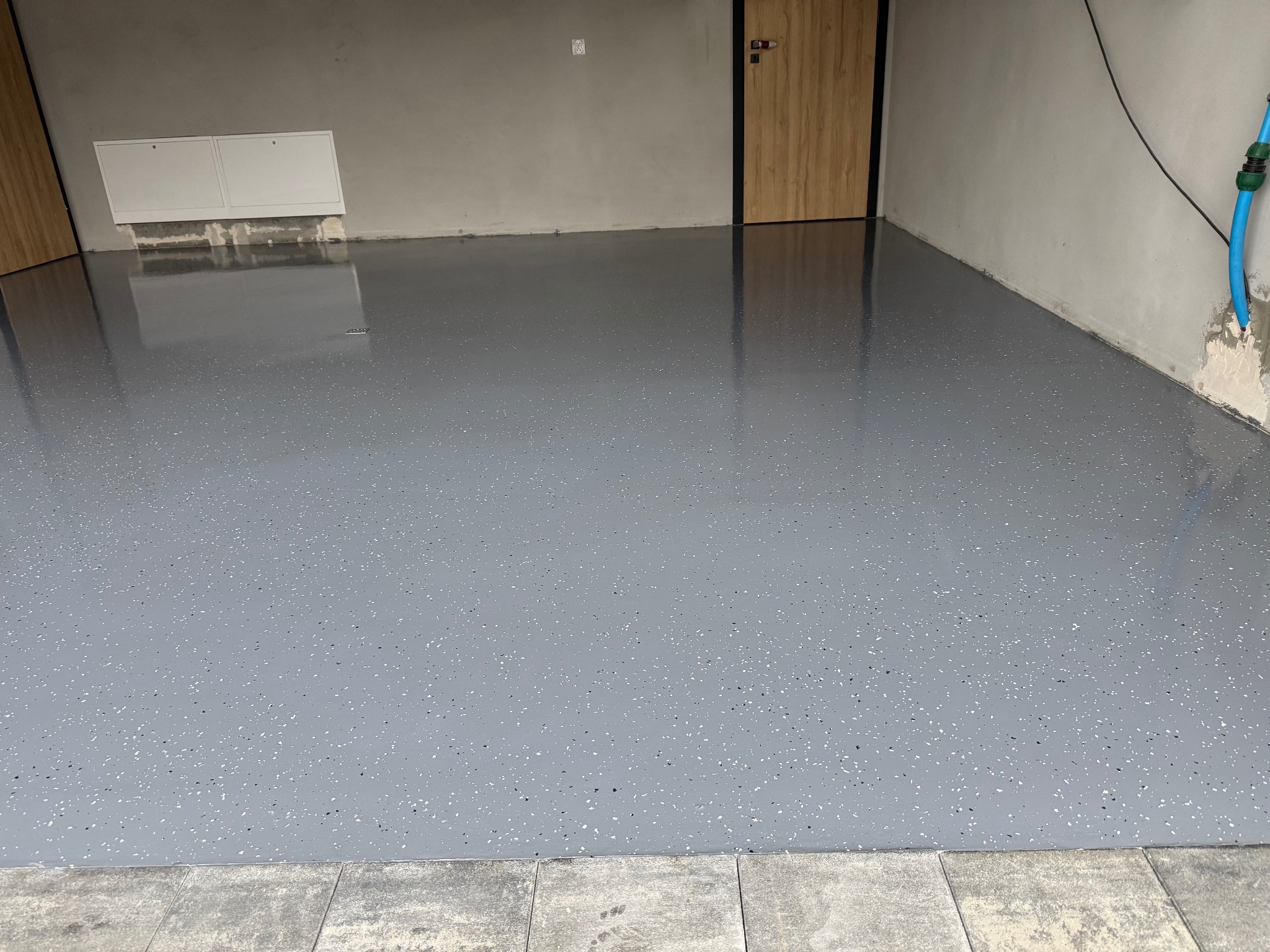 Szara, błyszcząca podłoga z drobnymi, ciemnymi i jasnymi plamkami w garażu lub pomieszczeniu gospodarczym, widoczne drzwi i ściany w tle.