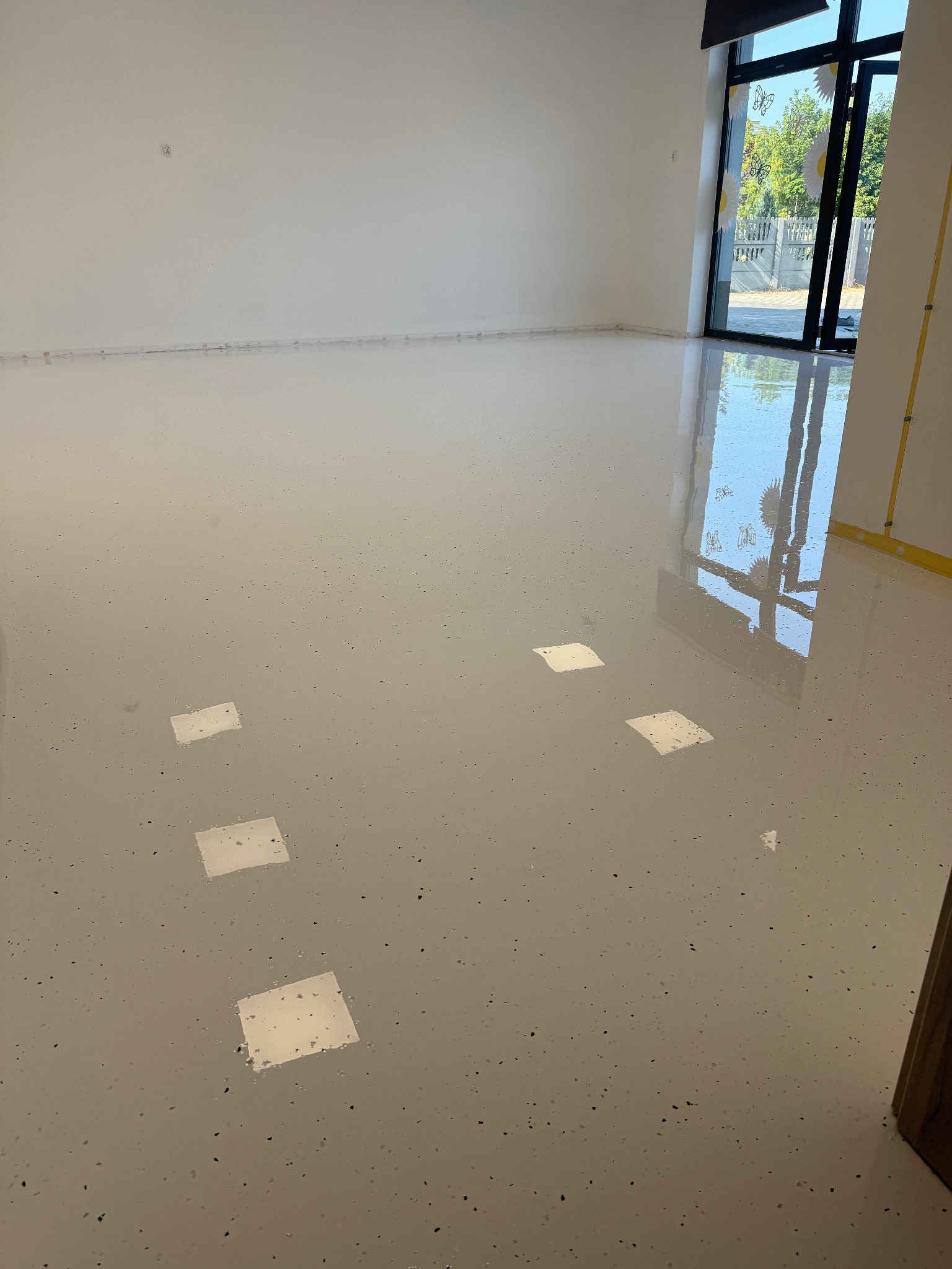 Jasna, błyszcząca podłoga z drobnymi, ciemnymi inkluzjami; okno z widocznymi dekoracjami i odbiciami.