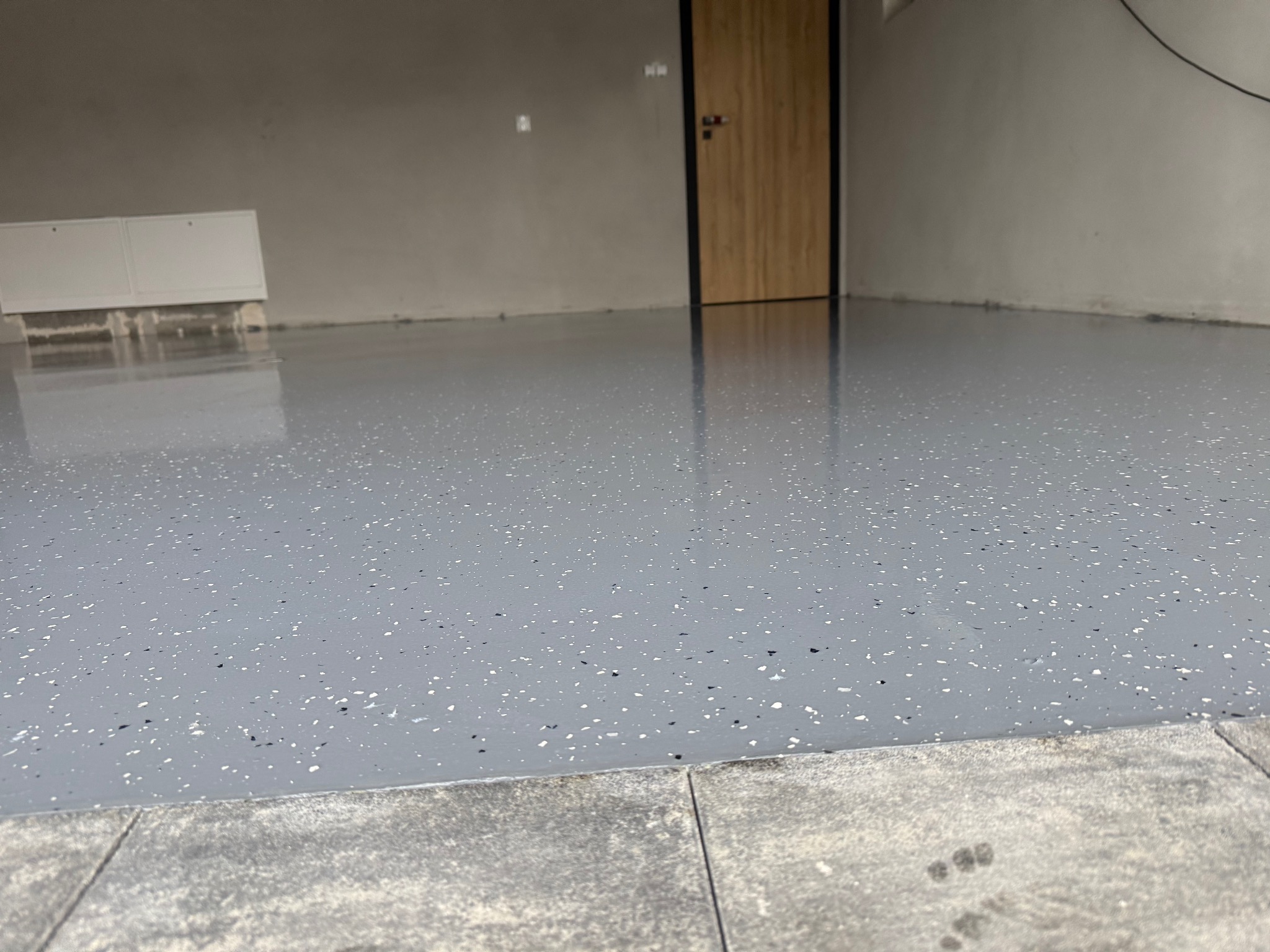 Szara, błyszcząca podłoga z drobnymi, czarnymi i białymi plamkami, widoczna przy drzwiach z drewnianą okleiną i szarych ścianach.