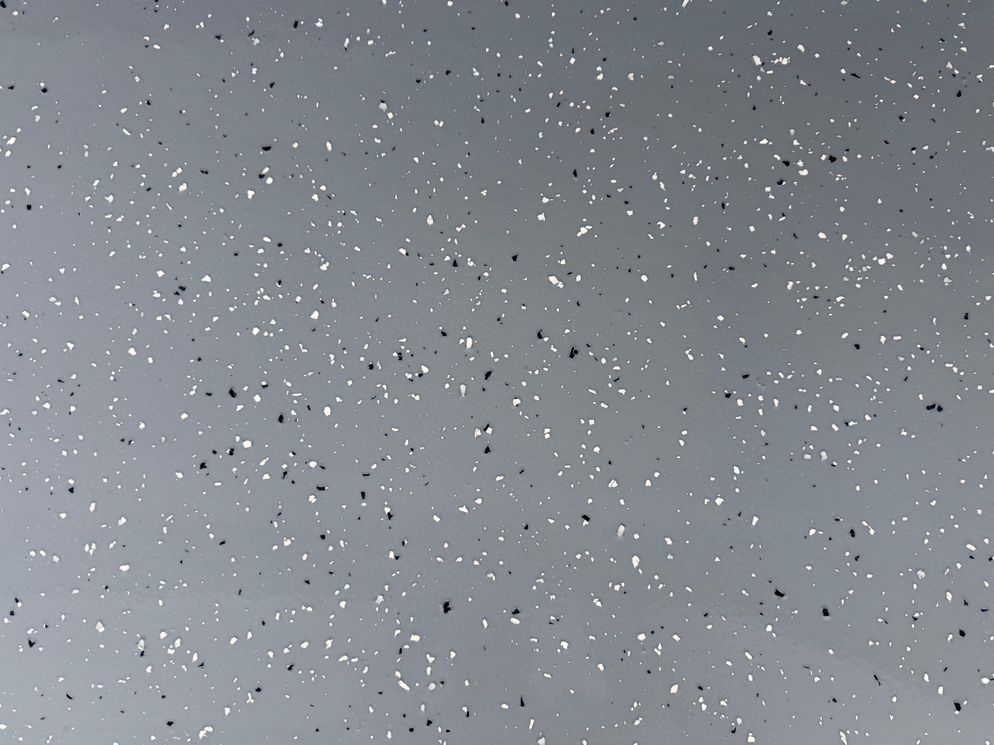 Szara posadzka żywiczna z drobnymi, nieregularnymi, czarnymi i białymi wtrąceniami, tworzącymi wzór przypominający lastryko. Powierzchnia gładka, jednolita, bez widocznych łączeń.