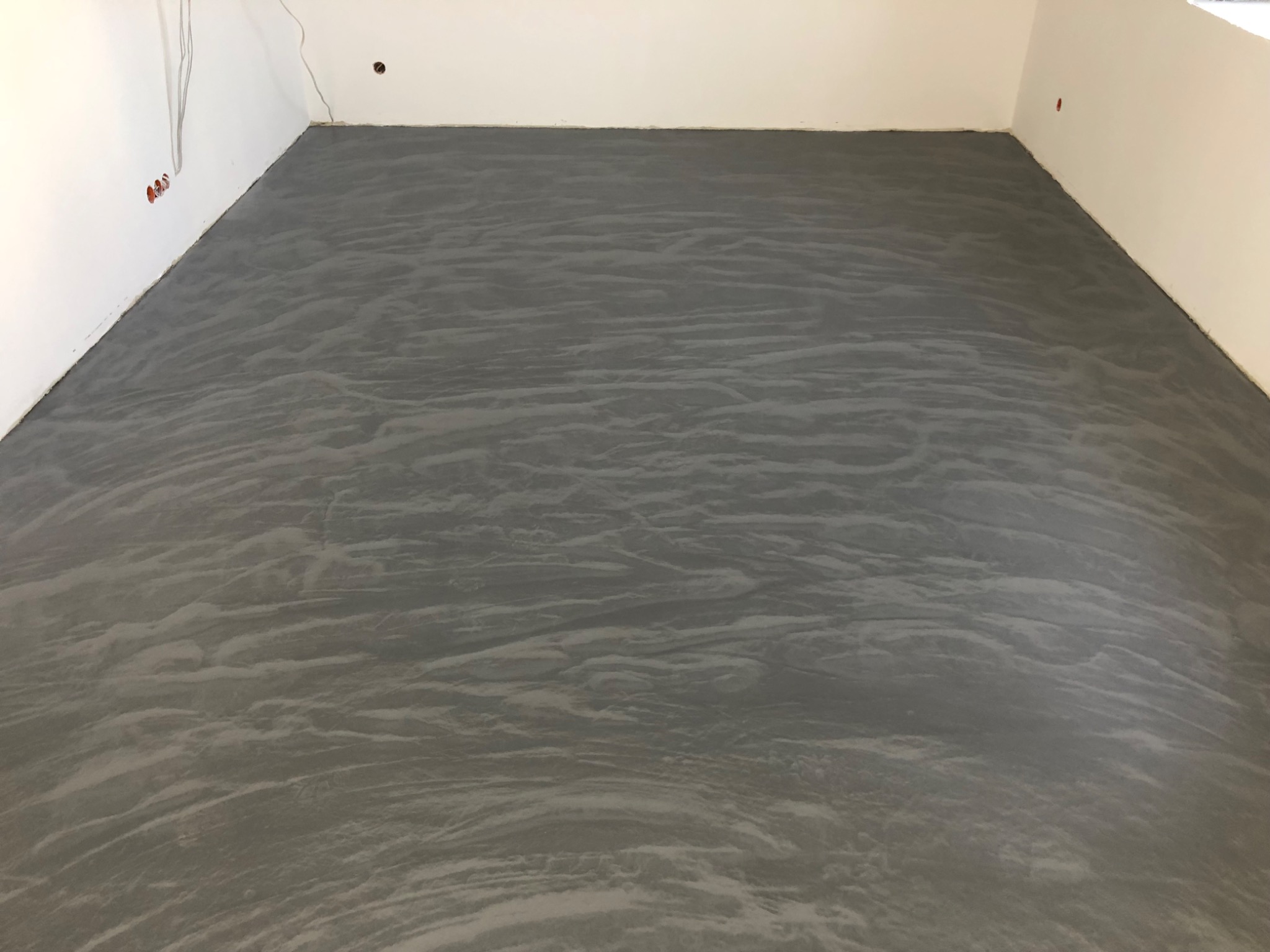 Szara, wzorzysta posadzka w pustym pomieszczeniu z białymi ścianami i widocznymi przewodami elektrycznymi.