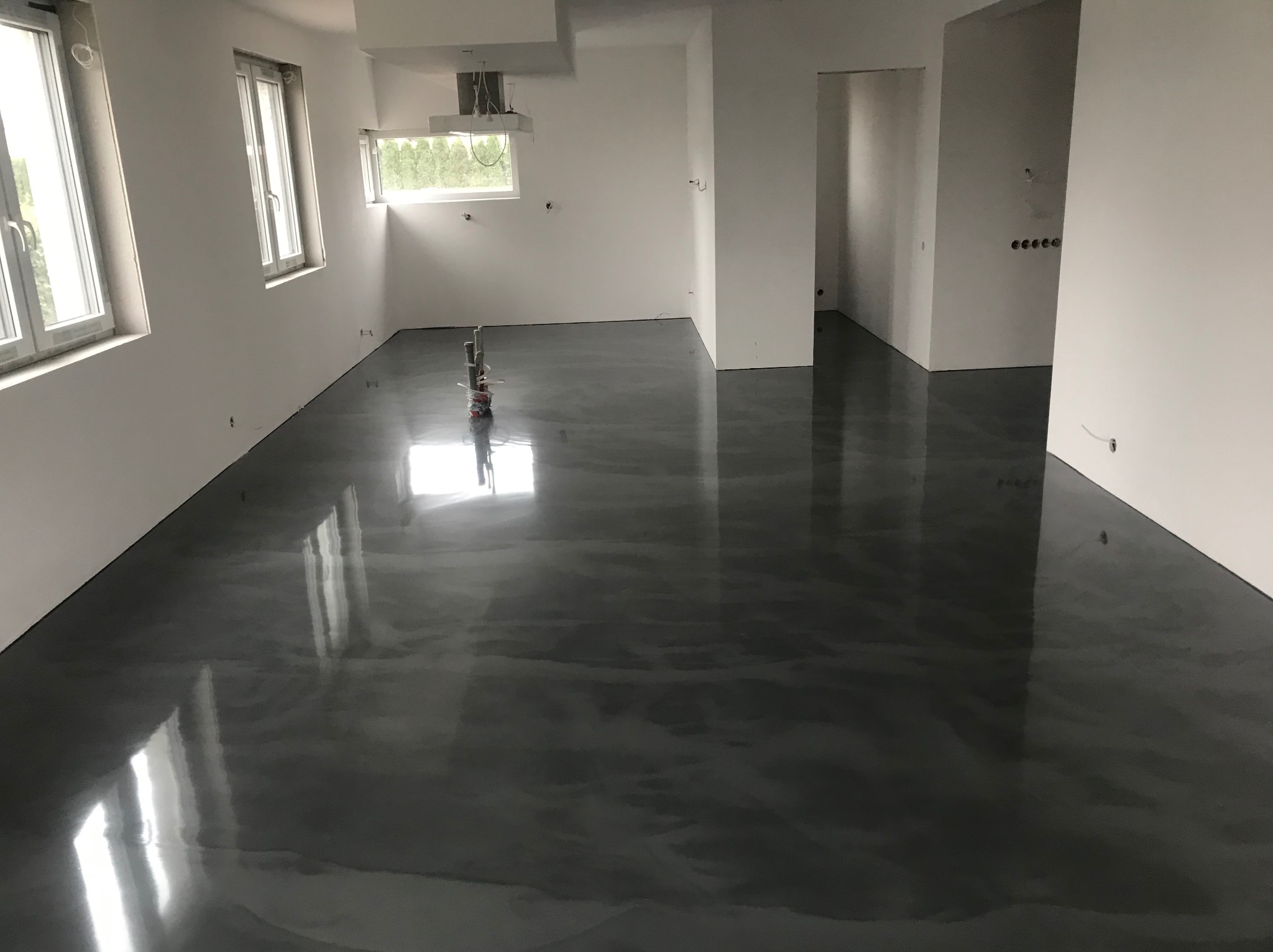 Świeżo wylana, błyszcząca, ciemnoszara posadzka w pustym, jasnym pomieszczeniu z oknami i widocznymi puszkami elektrycznymi w ścianie.