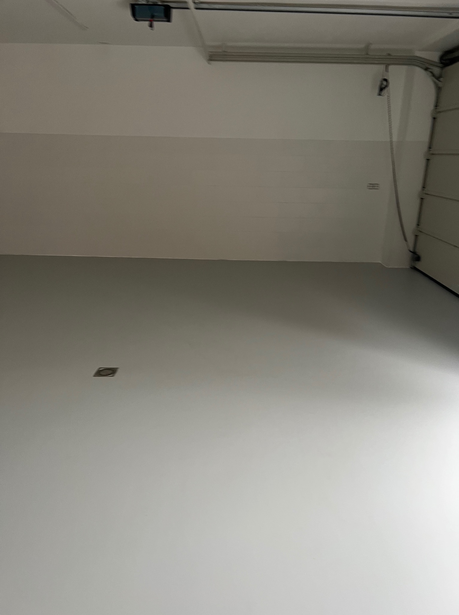 Jasnoszara, gładka posadzka w garażu z otwartą bramą segmentową po prawej stronie i kratką wentylacyjną w podłodze.