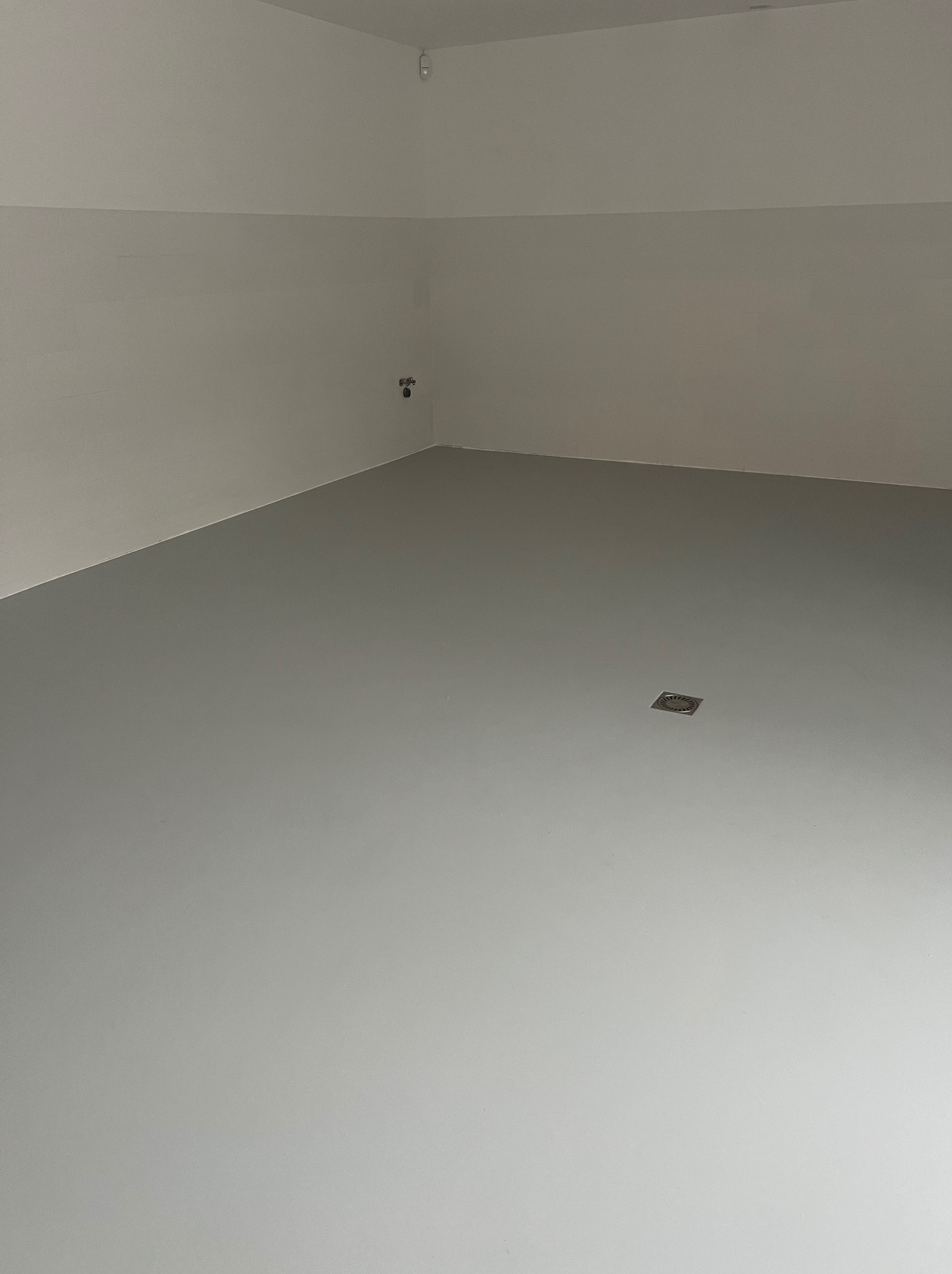 Świeżo wylana, gładka podłoga w jasnoszarym odcieniu, z widocznym odpływem liniowym w rogu pomieszczenia i instalacją na ścianie.