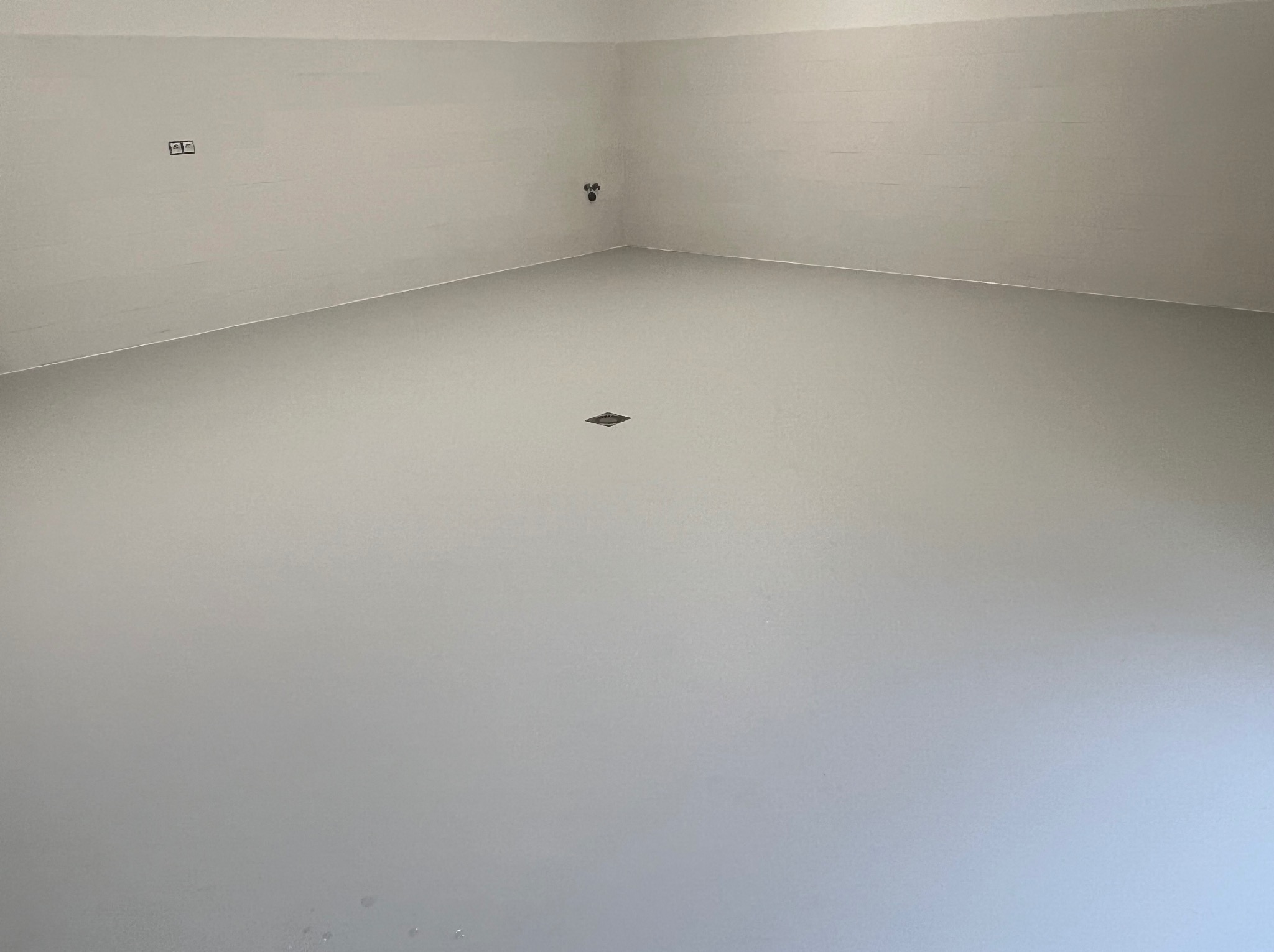 Jasnoszara, jednolita powierzchnia podłogi w pustym pomieszczeniu z odpływem podłogowym i instalacją na ścianie.