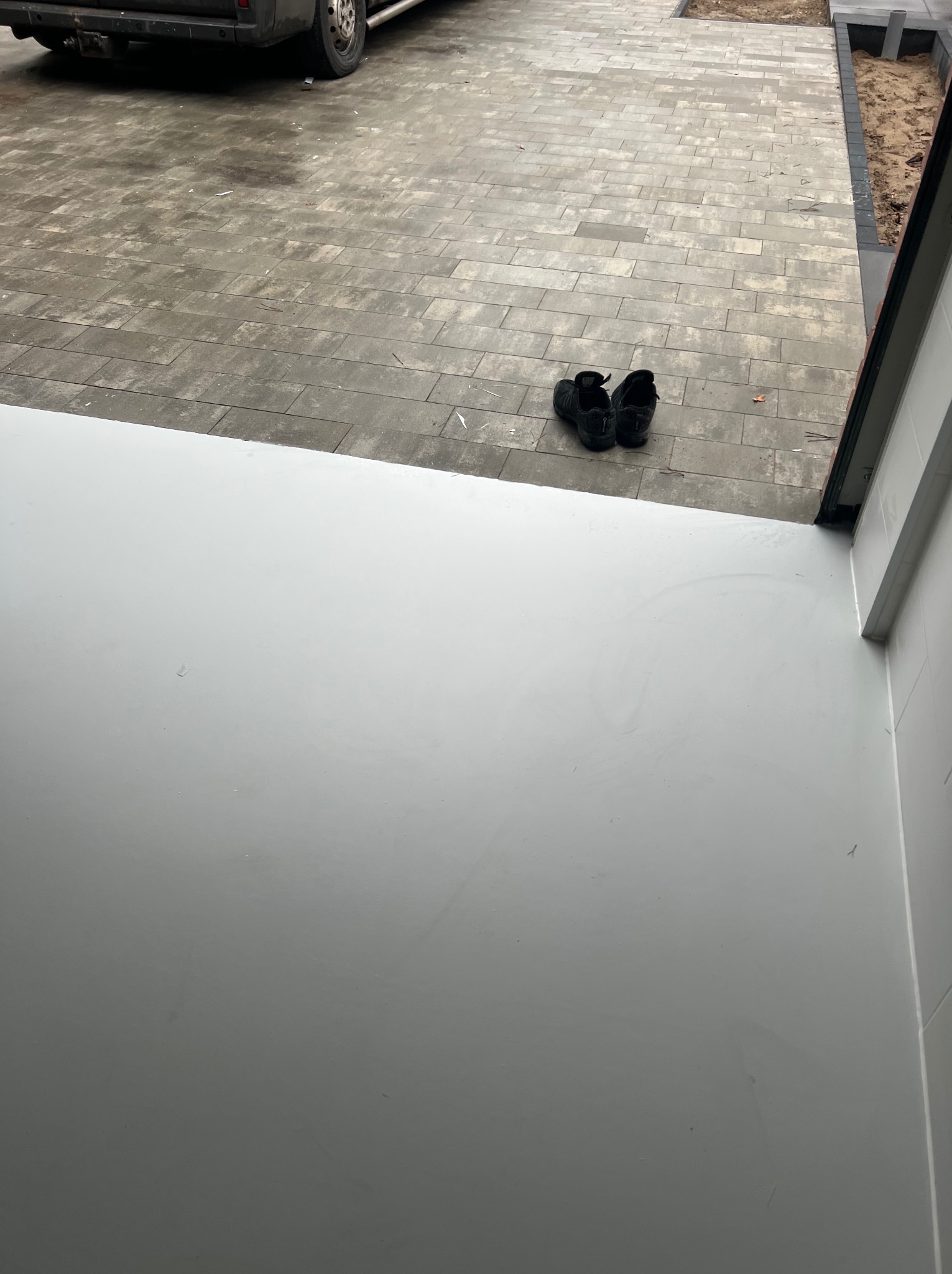 Świeżo wylana, jasnoszara posadzka w garażu z parą czarnych butów roboczych na zewnątrz, na kostce brukowej.